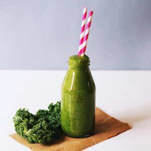 Green protein smoothie in a glass bottle with veggies on side on a white table